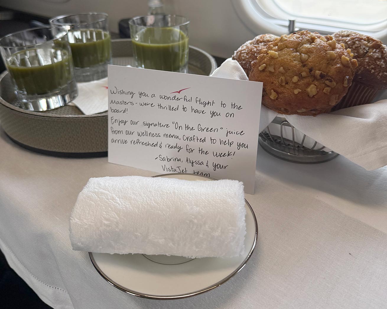 A rolled white towel on a plate, a card with a welcome message, two glasses of green juice, and two muffins on a tray are arranged on a white tablecloth next to an airplane window—first-class service fit for the Masters or even Gary Player himself.