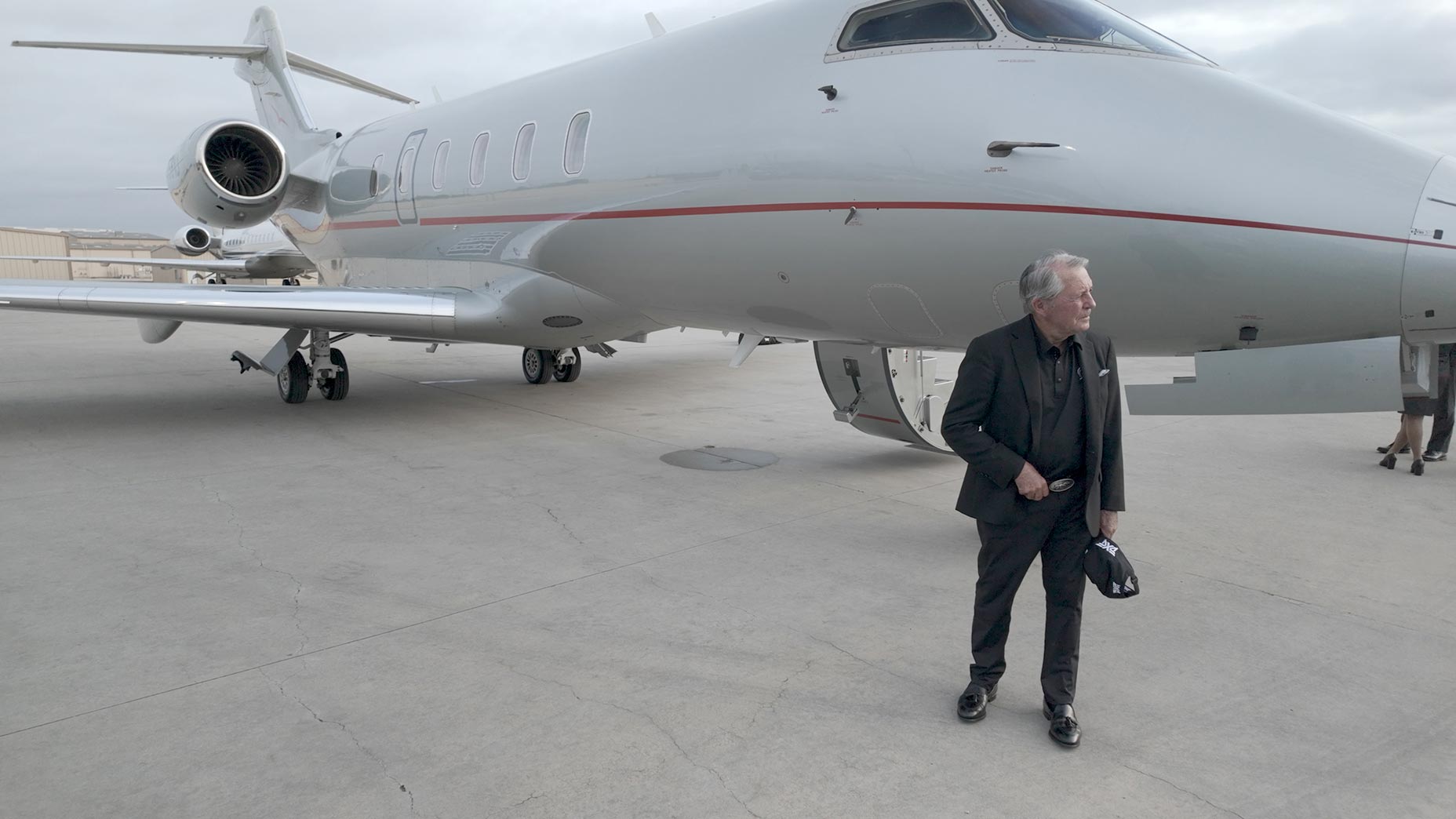 A man in a black suit stands on an airport tarmac next to a private jet, holding a black cap—emblazoned with the Masters logo—in his hand. The overcast weather sets the scene as another’s legs appear near the jet’s stairs, reminiscent of Gary Player’s travels.