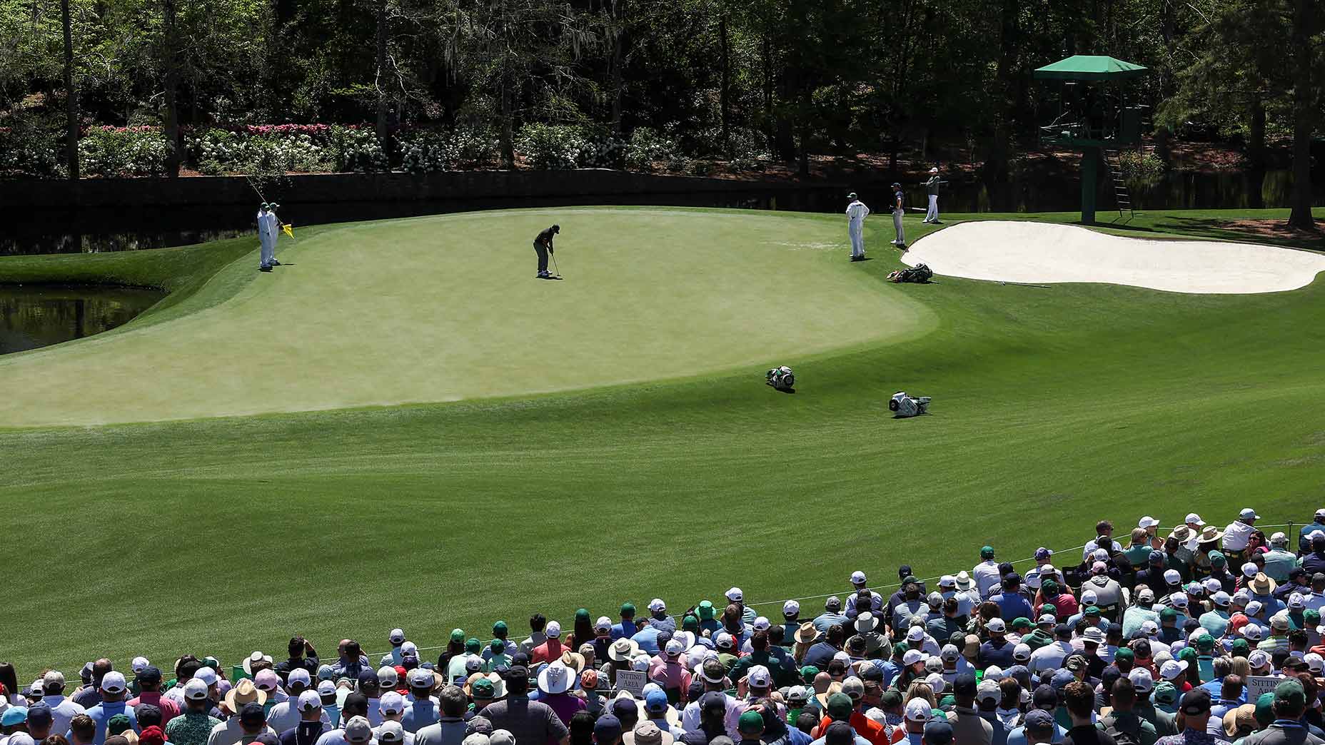 A good player prepares to putt on a green surrounded by sand traps at Augusta, while caddies and spectators watch. A large crowd sits in the foreground under sunny skies at this professional golf tournament.