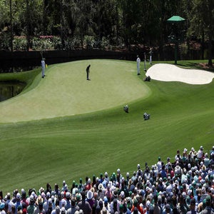 A good player prepares to putt on a green surrounded by sand traps at Augusta, while caddies and spectators watch. A large crowd sits in the foreground under sunny skies at this professional golf tournament.