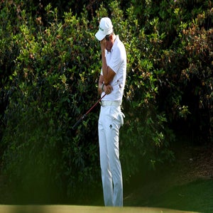 Haotong Li dressed in white stands on the green with his head bowed and hand on his face, appearing disappointed, at the Masters. Lush green bushes fill the background.