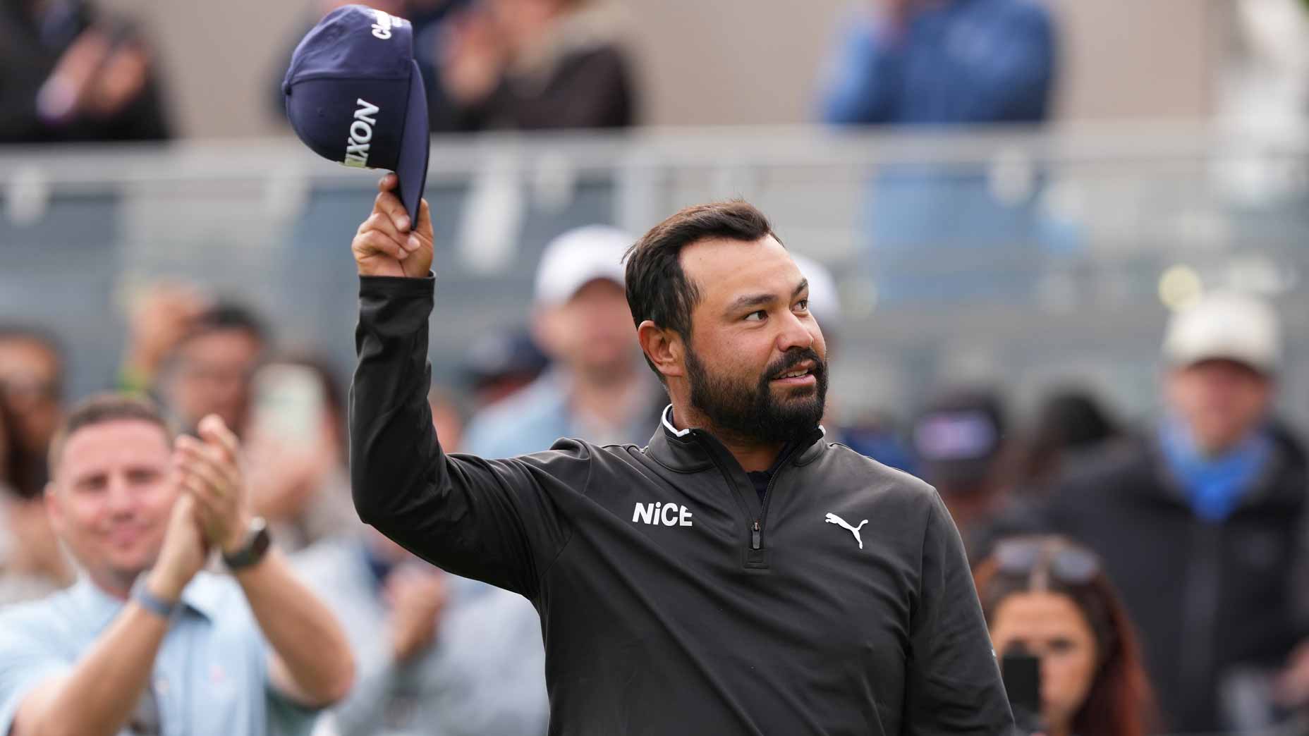 J.J. Spaun, wearing a black jacket and holding a cap in the air, stands in front of a cheering crowd at the Valero Texas Open, celebrating his victory and Masters qualification.