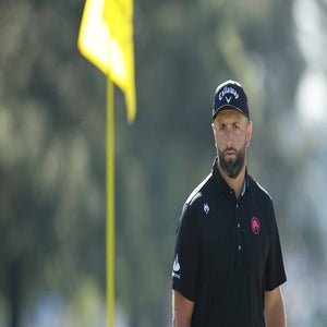A male golfer in a black shirt and cap stands on a golf course near a flagstick, looking focused; trees and a blurred yellow flag are visible in the background.