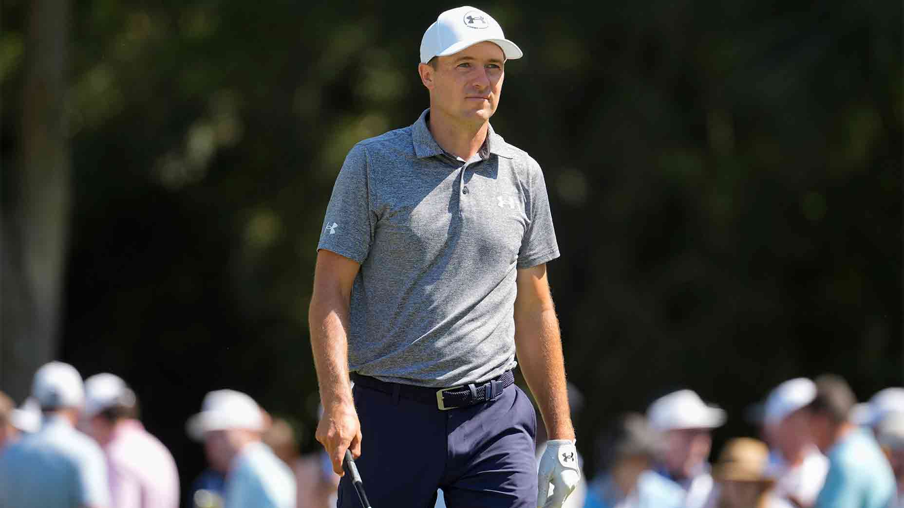 A male golfer, resembling Spieth, wears a blue Under Armour shirt, white cap, and navy pants as he stands on a golf course holding a club, with spectators blurred in the background.
