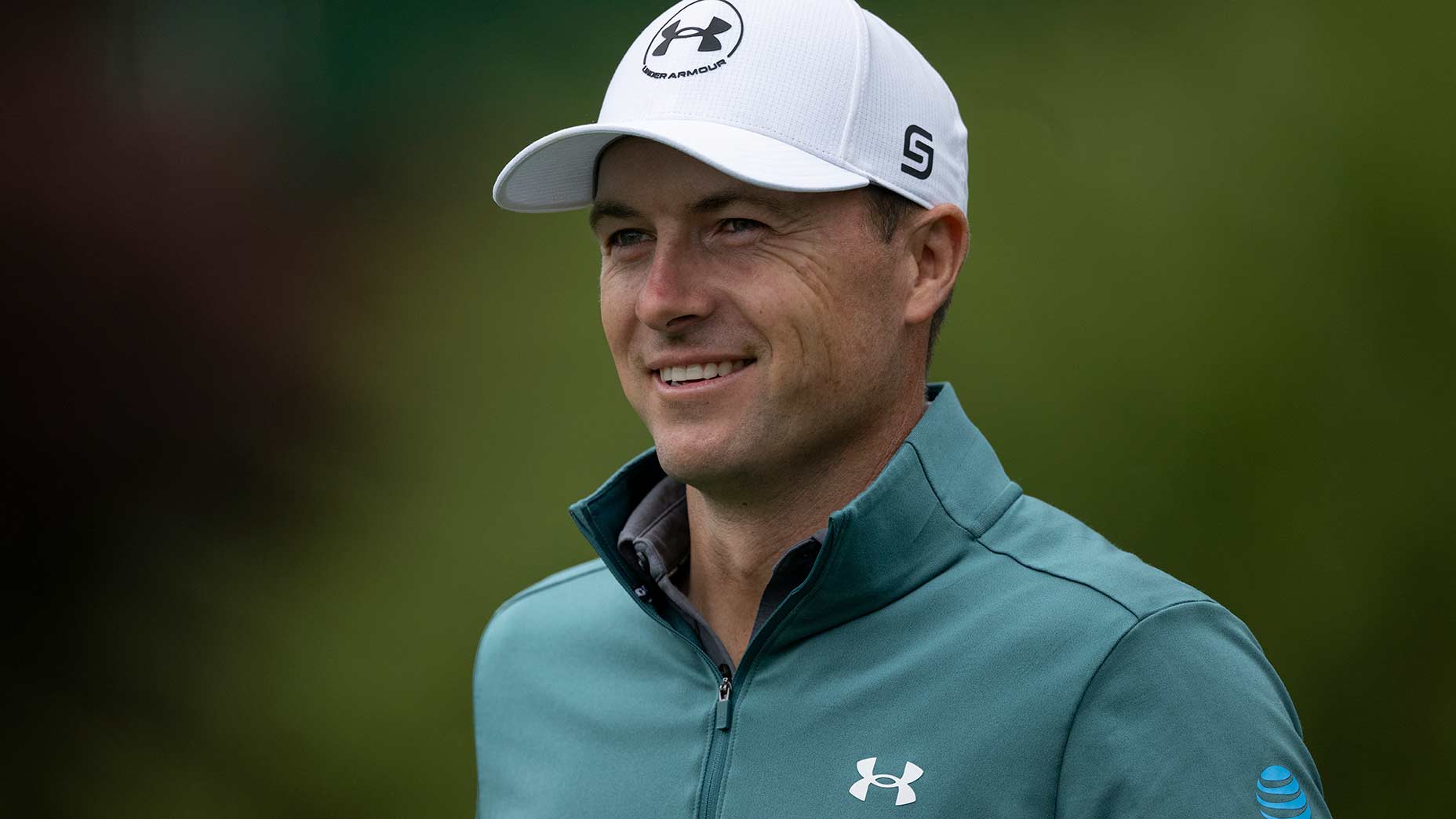 A smiling man wearing a white cap and teal Under Armour jacket stands outdoors against a blurred green background.