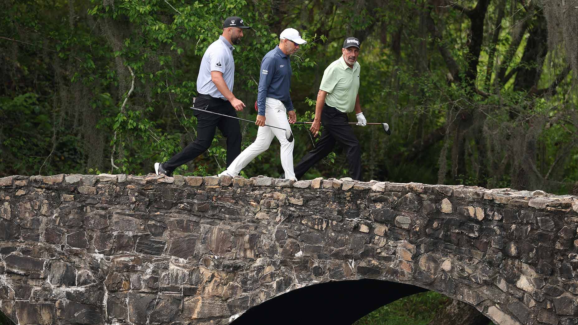 Three golfers walk together across a stone bridge surrounded by greenery, each holding golf clubs and dressed in golf attire, during a round of golf.