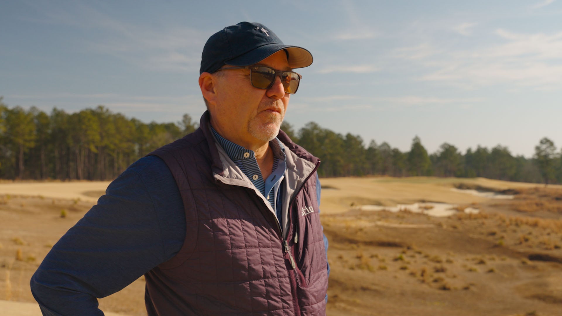 A man wearing sunglasses, a baseball cap, and a quilted jacket stands outdoors on a sunny day at a tree farm, with sandy terrain and pine trees in the background.