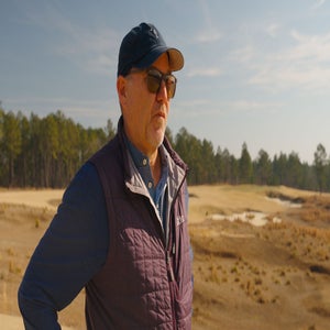 A man wearing sunglasses, a baseball cap, and a quilted jacket stands outdoors on a sunny day at a tree farm, with sandy terrain and pine trees in the background.