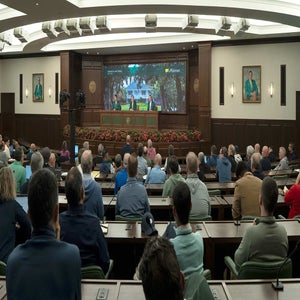 A large group of people sits facing a panel of four speakers at the front of a conference room, with portraits on the walls and a screen displaying the Masters golf tournament logo and an outdoor scene.