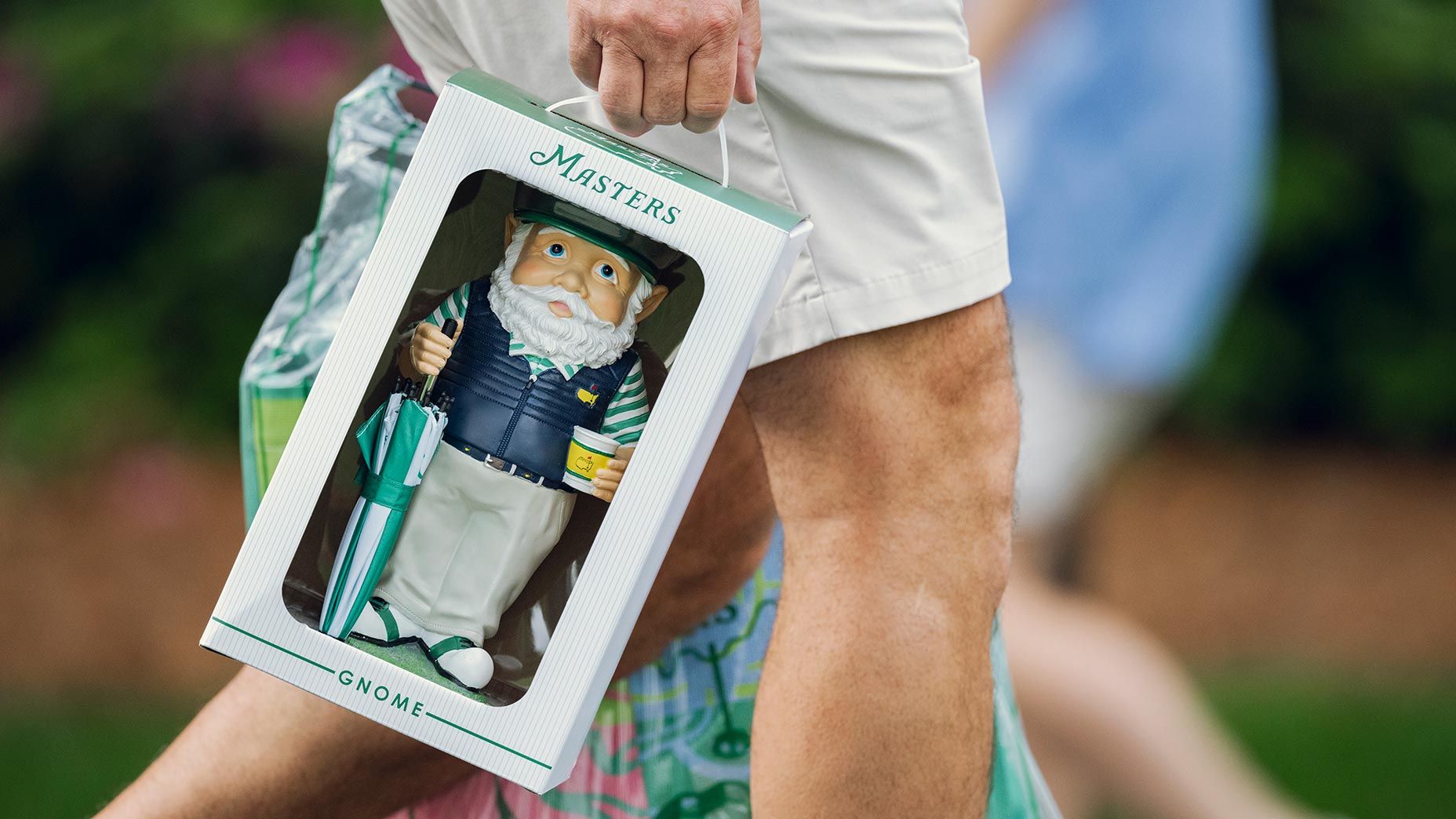 A person holds a boxed Masters gnome figurine, featuring a bearded gnome in golf attire holding a cup and umbrella, while walking outdoors.