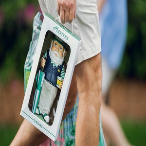 A person holds a boxed Masters gnome figurine, featuring a bearded gnome in golf attire holding a cup and umbrella, while walking outdoors.