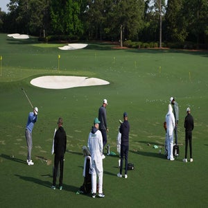Golfers practice their swings on a green driving range, with sand bunkers and yellow flags in the background. Several caddies and coaches stand nearby observing and assisting. Trees border the range under a clear sky.