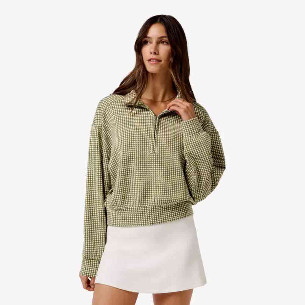 A woman wearing a long-sleeved green and white checkered sweater and a white skirt stands against a plain light background, looking slightly to the side – an elegant look perfect for Mother's Day.