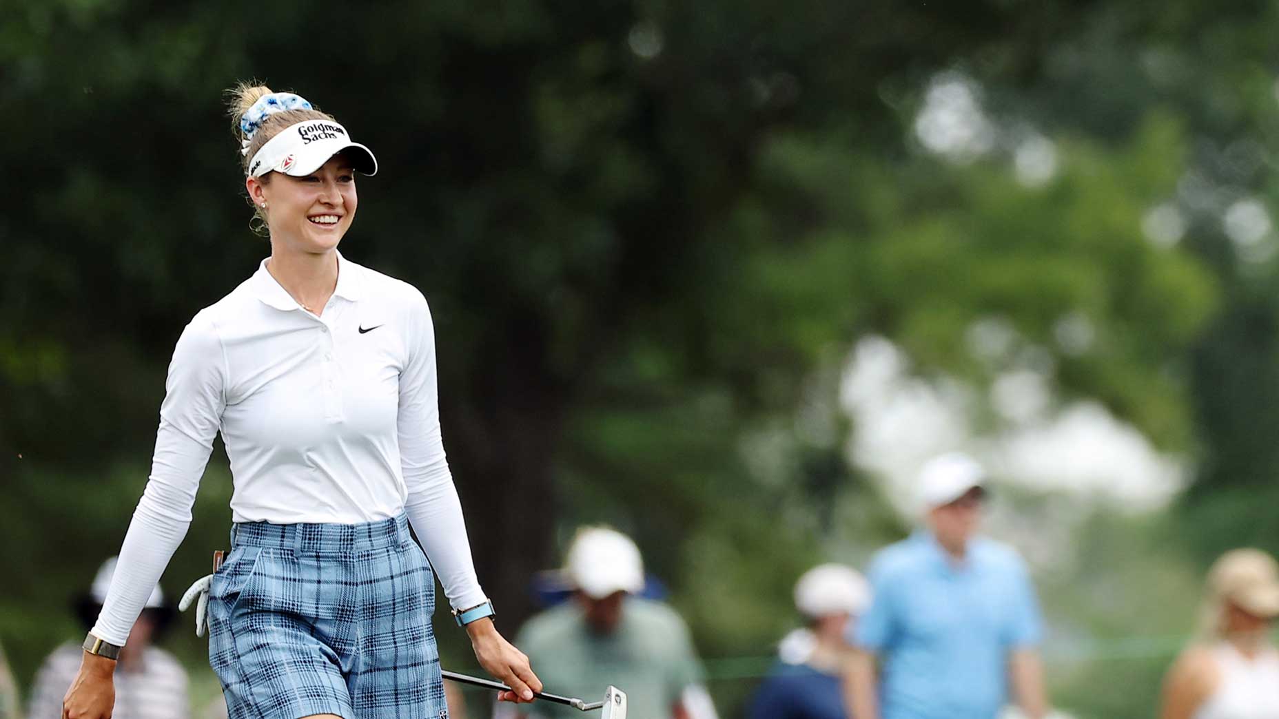Nelly Korda in a white long-sleeve shirt and blue plaid pants smiles while holding a putter on the golf course during the 2026 Chevron Championship, with blurred trees and people in the background.