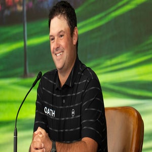 A man wearing a black striped polo shirt with the word OATH on it sits smiling at a table in front of a microphone, with a green, outdoor background behind him&mdash;reminiscent of Patrick Reed at the Masters.