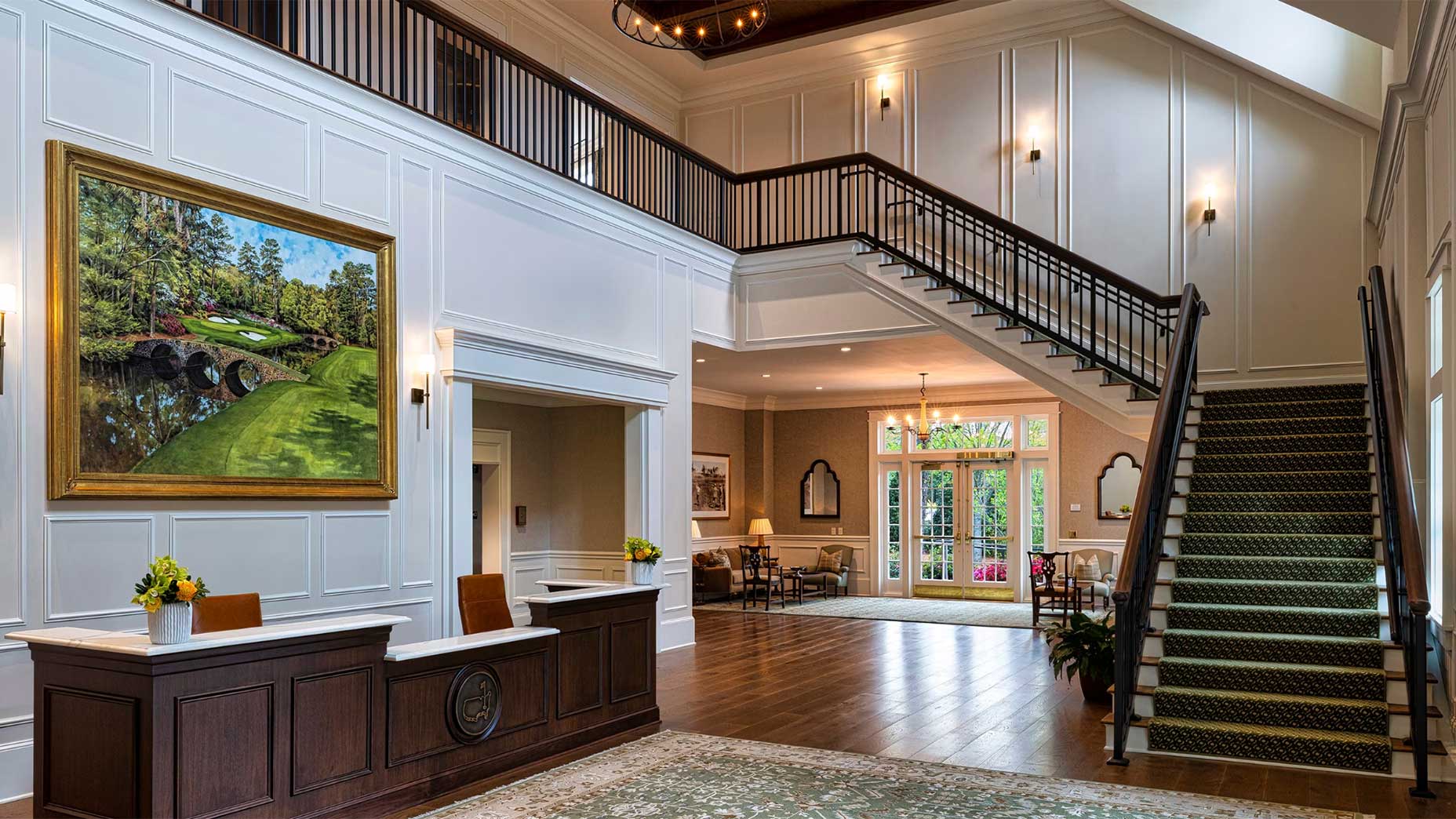 Elegant lobby with a dark wood reception desk, large golf course painting, patterned rug, and grand staircase with green carpet. The space features white paneled walls, large windows, and comfortable seating.