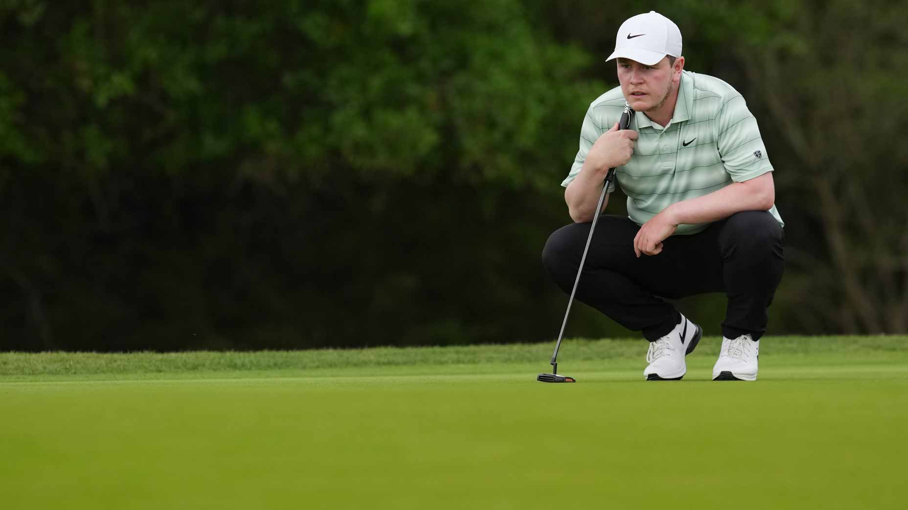 A golfer in a white cap and striped shirt crouches on the green, focusing on lining up a putt, with trees in the background.