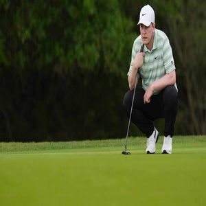 A golfer in a white cap and striped shirt crouches on the green, focusing on lining up a putt, with trees in the background.