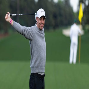 Rory McIlroy in a gray sweater and black cap stretches with a golf club on a green course, smiling, while another person stands near a yellow flag in the background.