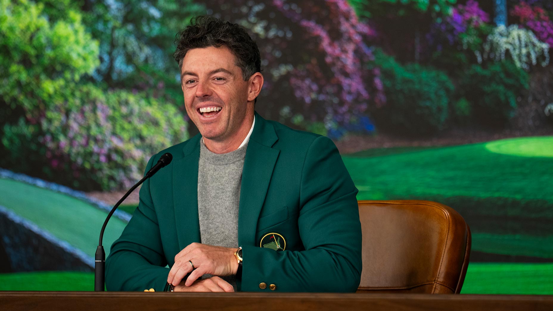 A man in a green jacket sits at a desk with a microphone, smiling during a press conference. The background shows a vibrant golf course scene, hinting at the Masters Champions Dinner, with colorful flowers and greenery.