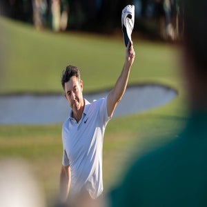 A golfer wearing a light-colored polo shirt raises his cap in the air while standing on a Masters golf course near a sand bunker, acknowledging the crowd.