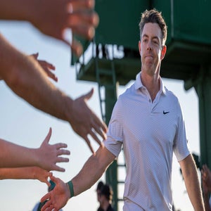Rory McIlroy in a white polo shirt walks by and high-fives fans reaching out their hands, smiling as he passes by under a clear sky at the Masters.