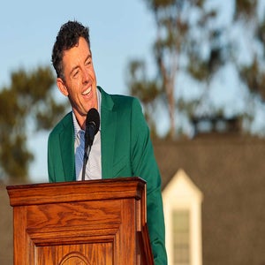 A man wearing a green jacket smiles while speaking at a wooden podium outdoors, with a blurred building and trees in the background.