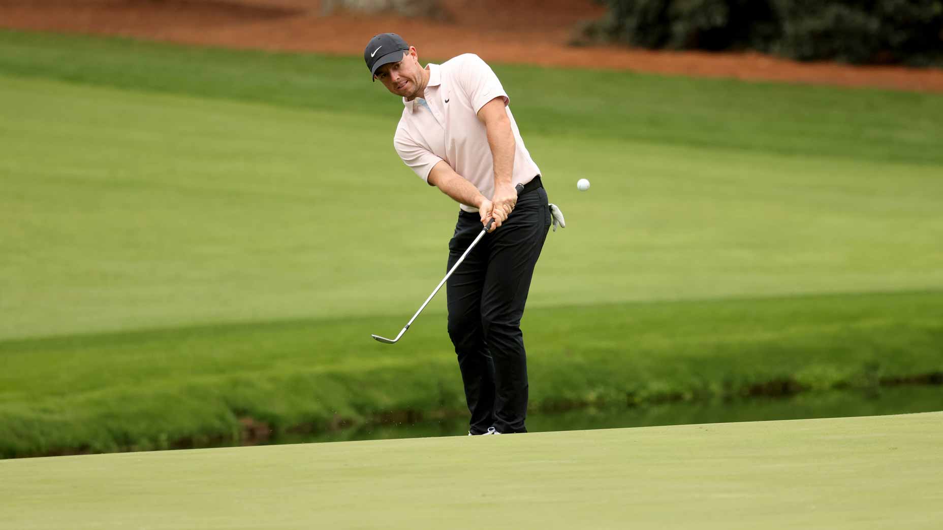 A golfer wearing a light pink shirt, black pants, and a dark cap is mid-swing, chipping a golf ball onto the green at a golf course with neatly trimmed grass and a hint of trees in the background.