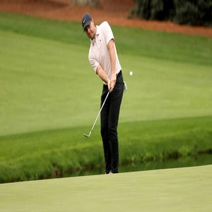 A golfer wearing a light pink shirt, black pants, and a dark cap is mid-swing, chipping a golf ball onto the green at a golf course with neatly trimmed grass and a hint of trees in the background.