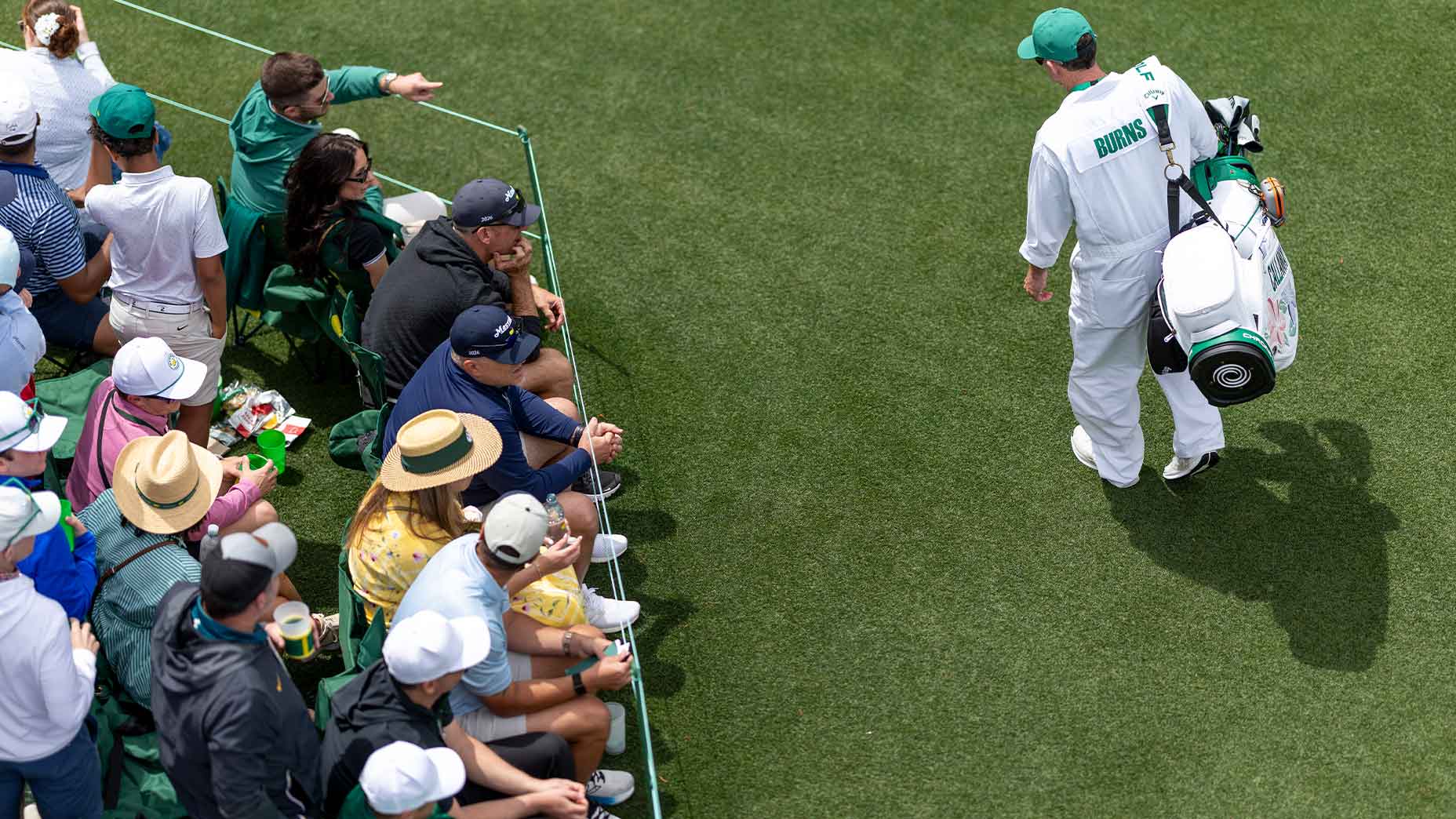 A golf caddie walks on a green field carrying a golf bag, while spectators in hats and casual clothing sit closely together along the edge, watching the action.