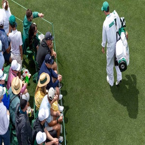 A golf caddie walks on a green field carrying a golf bag, while spectators in hats and casual clothing sit closely together along the edge, watching the action.