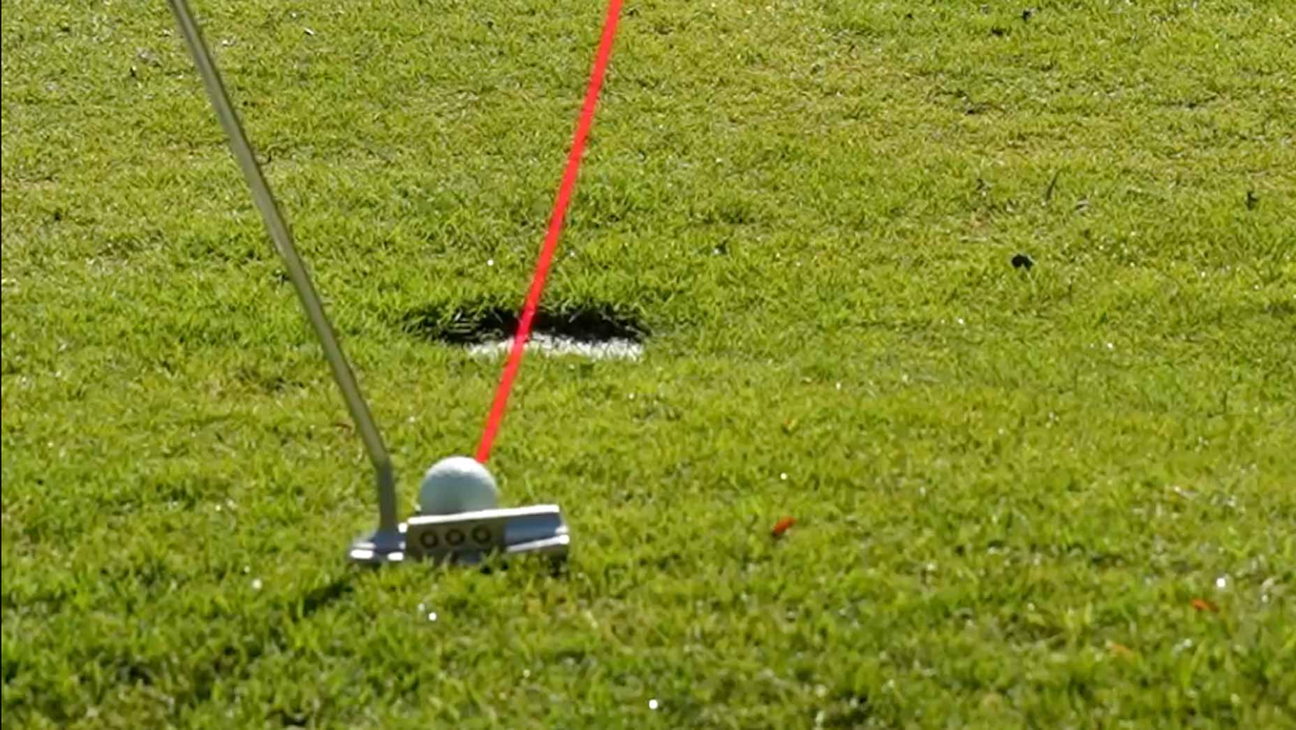 A golf putter is positioned behind a golf ball on green grass, aiming toward a hole. A red laser line projects from the putter, aligning the shot directly with the hole.