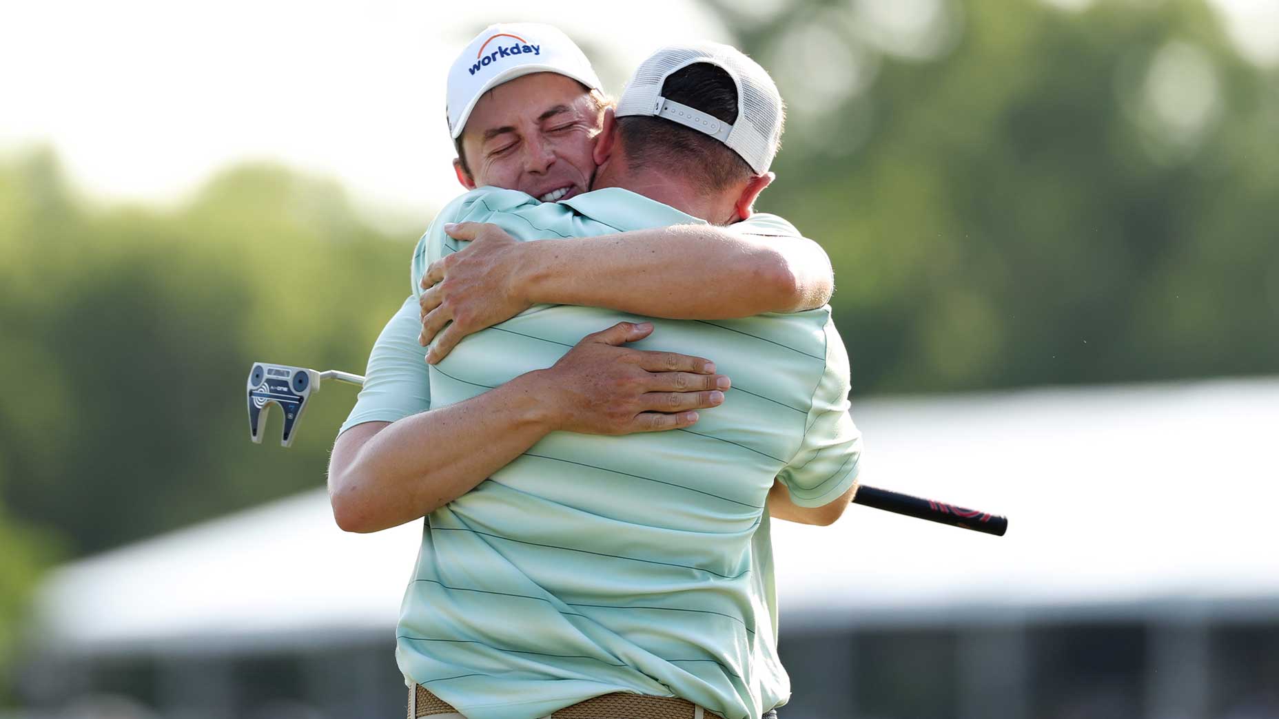 Alex and Matt Fitzpatrick embrace tightly on a golf course, one holding a golf club. The background is blurred with greenery and a structure visible—Matt Fitzpatrick celebrates on the PGA Tour.