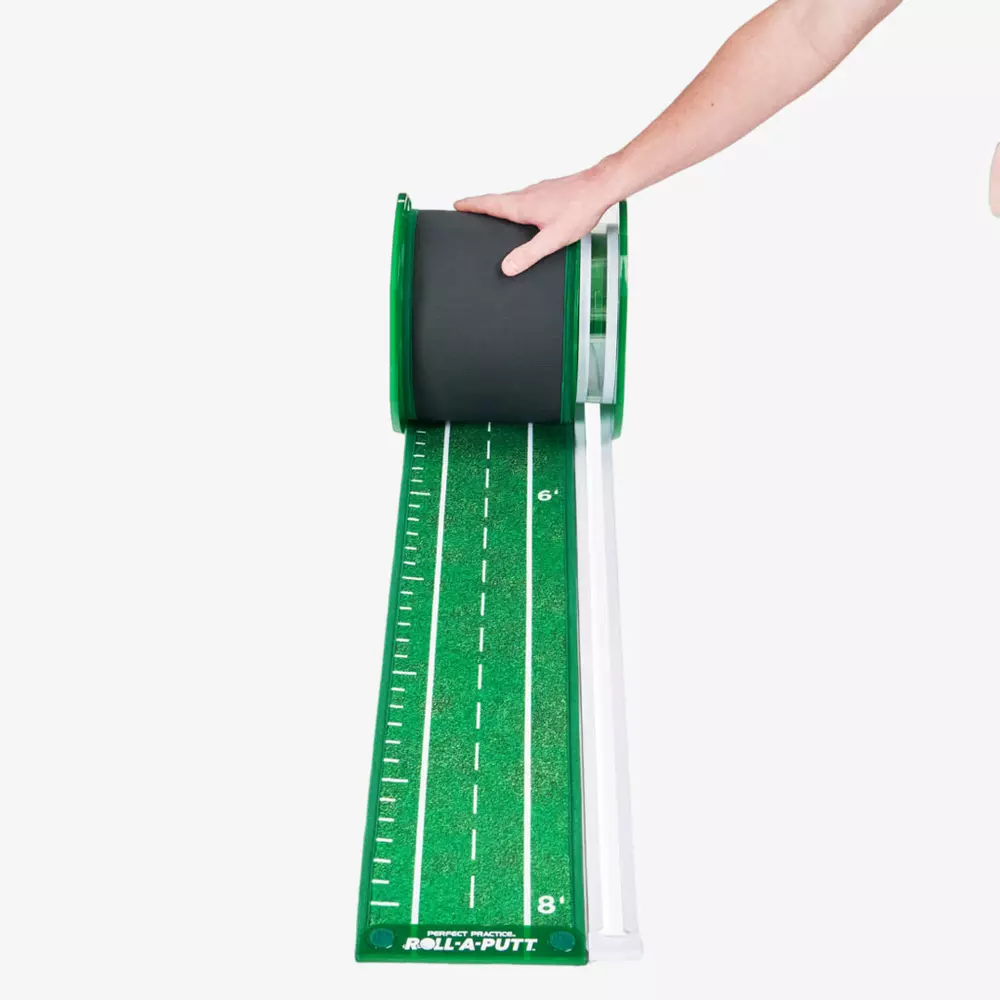 A person&rsquo;s hand unrolls a green putting mat with white distance markings and a simulated turf surface, resembling a miniature golf fairway, onto a white floor&mdash;one of the best deals under $50.