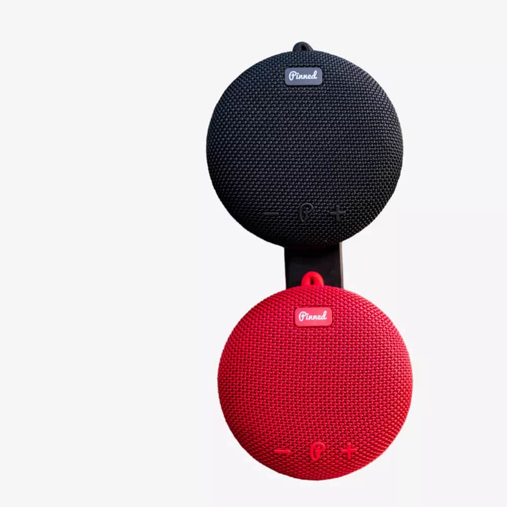 Two round, portable wireless speakers, one black and one red, are stacked vertically against a white background. Both feature fabric textures, control buttons, and the brand logo—perfect for those seeking the best deals under $50.