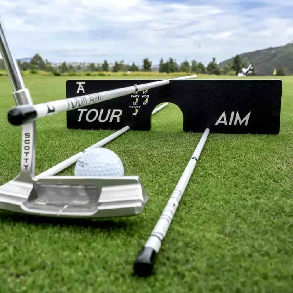 A golf putter, a golf ball, and two alignment rods are set up on grass with a black Tour Aim training aid in the background—an ideal setup for practicing golf alignment and finding the best deals under $50. Trees and hills can be seen in the distance.