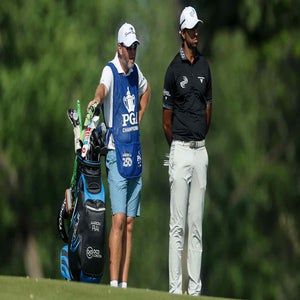 A golfer and his caddie stand on a golf course, both looking into the distance. The caddie leans on a golf bag filled with clubs fitted with inexpensive iron covers, while the golfer stands with arms crossed. Trees blur in the background.