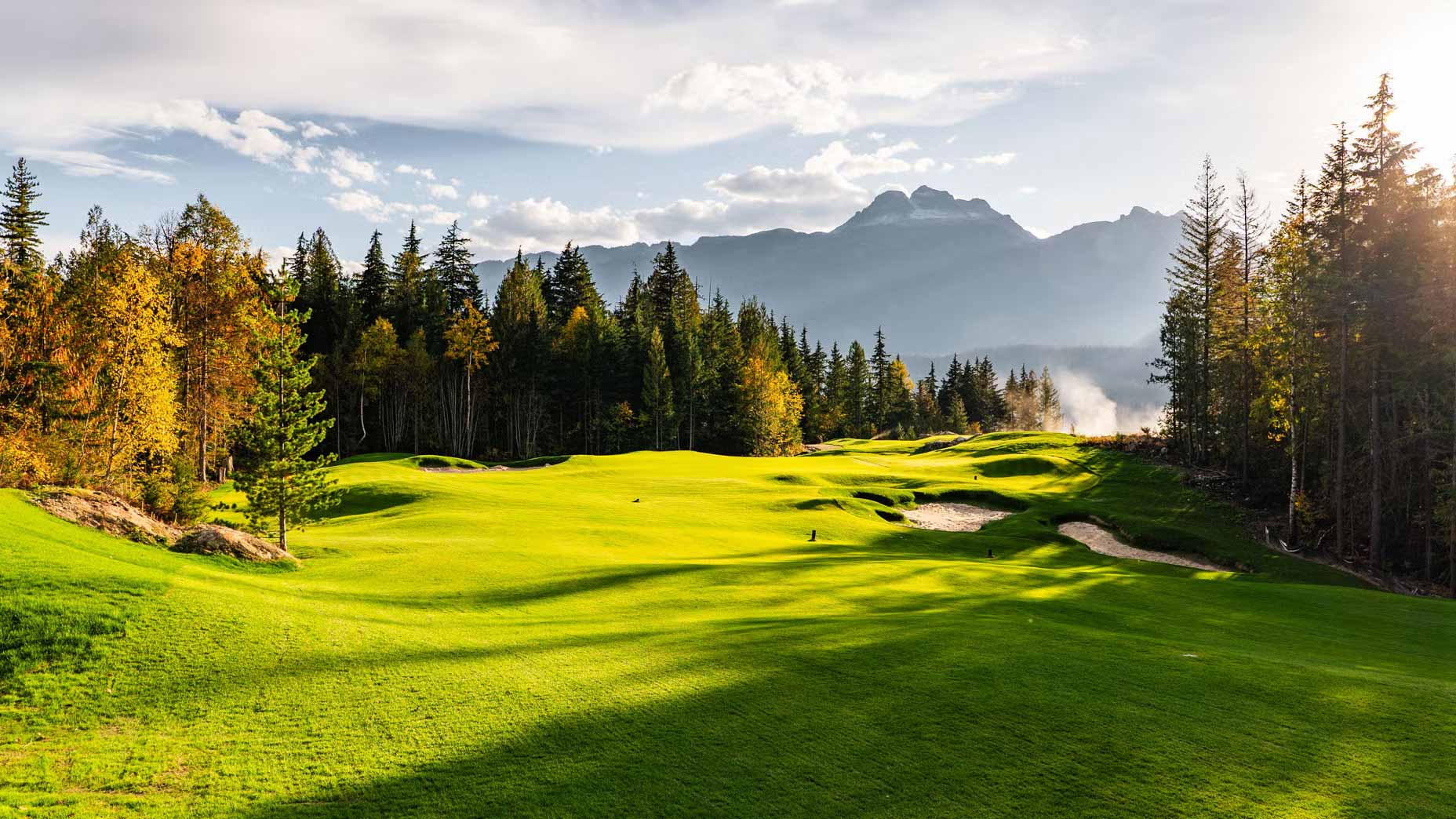 A lush green golf course at Cabot Revelstoke, surrounded by tall trees with autumn foliage, set against distant mountains under a partly cloudy sky, with sunlight casting long shadows on the grass.