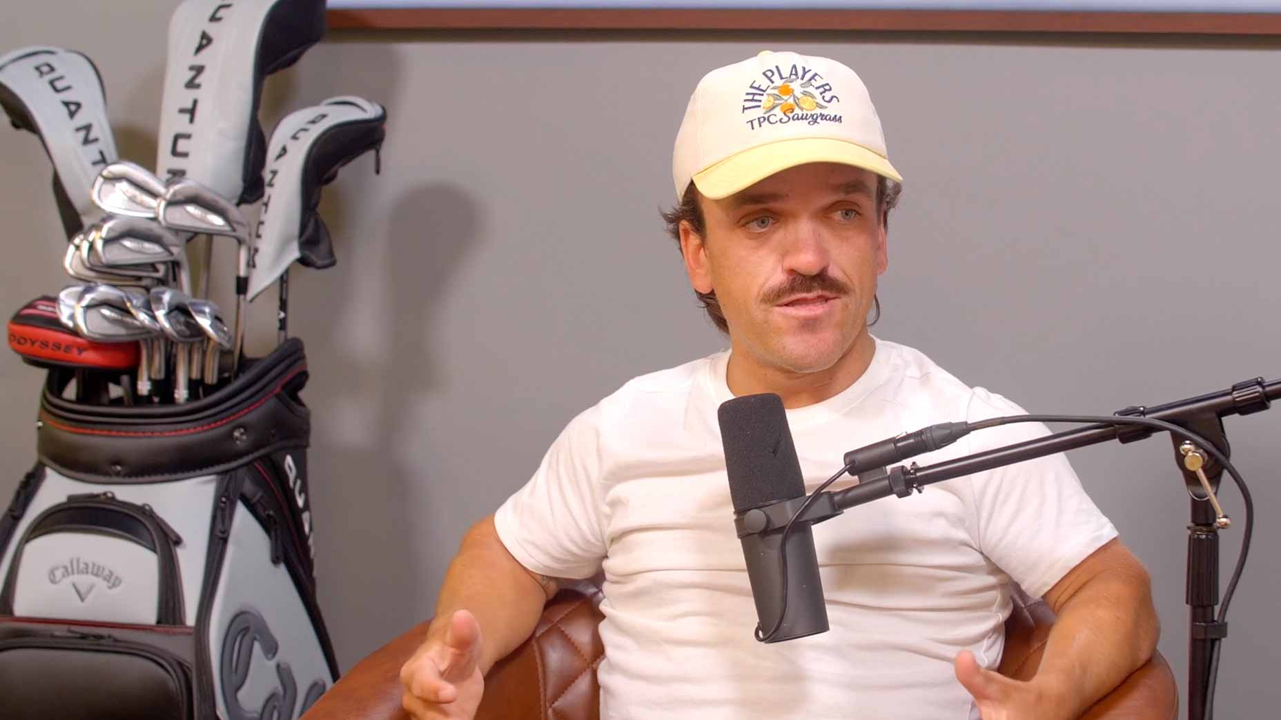 Chaz Bowker, dressed in a white t-shirt and cream hat, sits in a leather chair speaking into a microphone. Behind him is a golf bag with clubs.