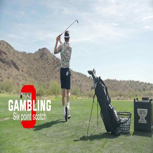 A golfer swings a club on a grassy course with desert hills in the background. Golf bag and basket of balls nearby. Text reads: GAMBLING Six point scotch, inspired by Drew Stoltz, with a large red G behind the word GAMBLING-game advantage.