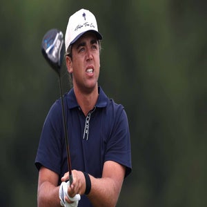 Garrick Higgo, wearing a navy shirt and white cap, watches his shot while holding a golf club against a blurred green background, unfazed by any costly late-arrival penalty.