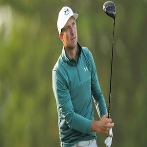 A male golfer wearing a white cap and teal long-sleeve shirt holds a driver after taking a shot, looking into the distance. The background is blurred greenery.