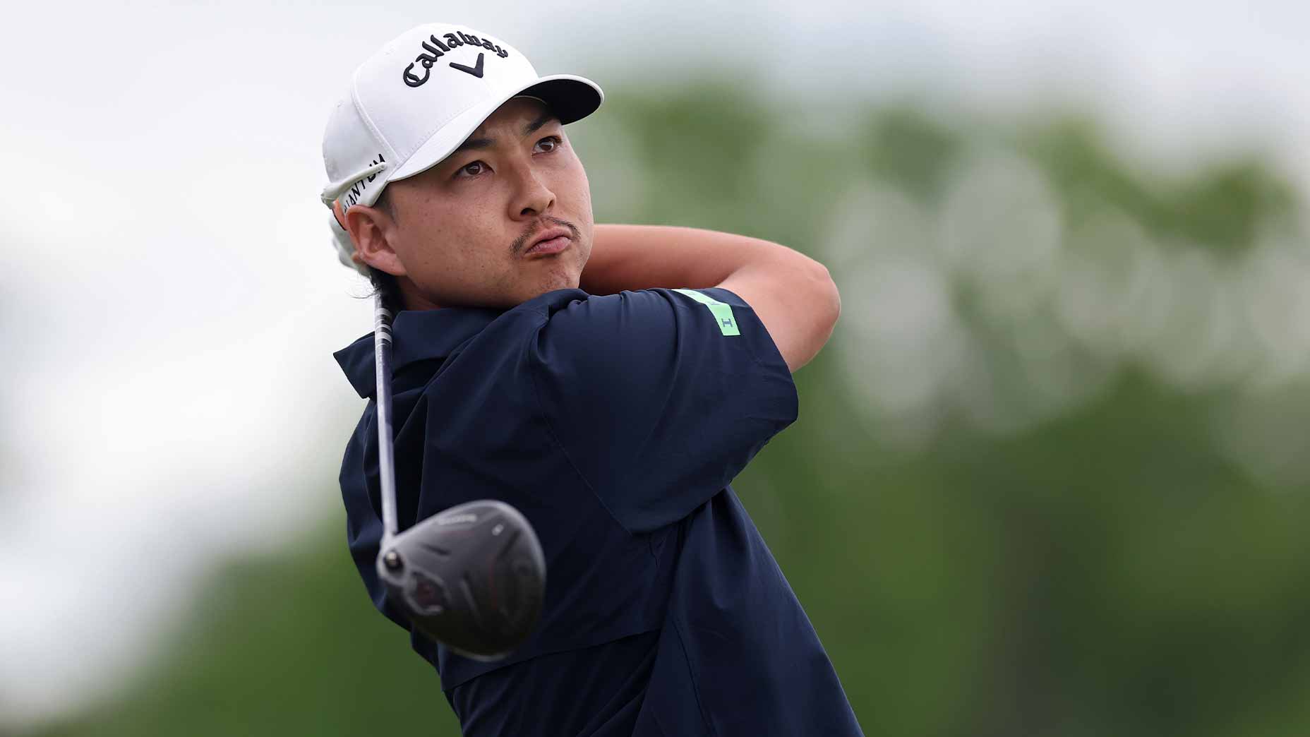 A golfer wearing a white cap and navy shirt follows through on a swing with his driver, evoking the intensity of the PGA Championship. The background is blurred, showing lush greenery beneath a cloudy sky.