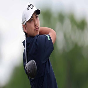 A golfer wearing a white cap and navy shirt follows through on a swing with his driver, evoking the intensity of the PGA Championship. The background is blurred, showing lush greenery beneath a cloudy sky.
