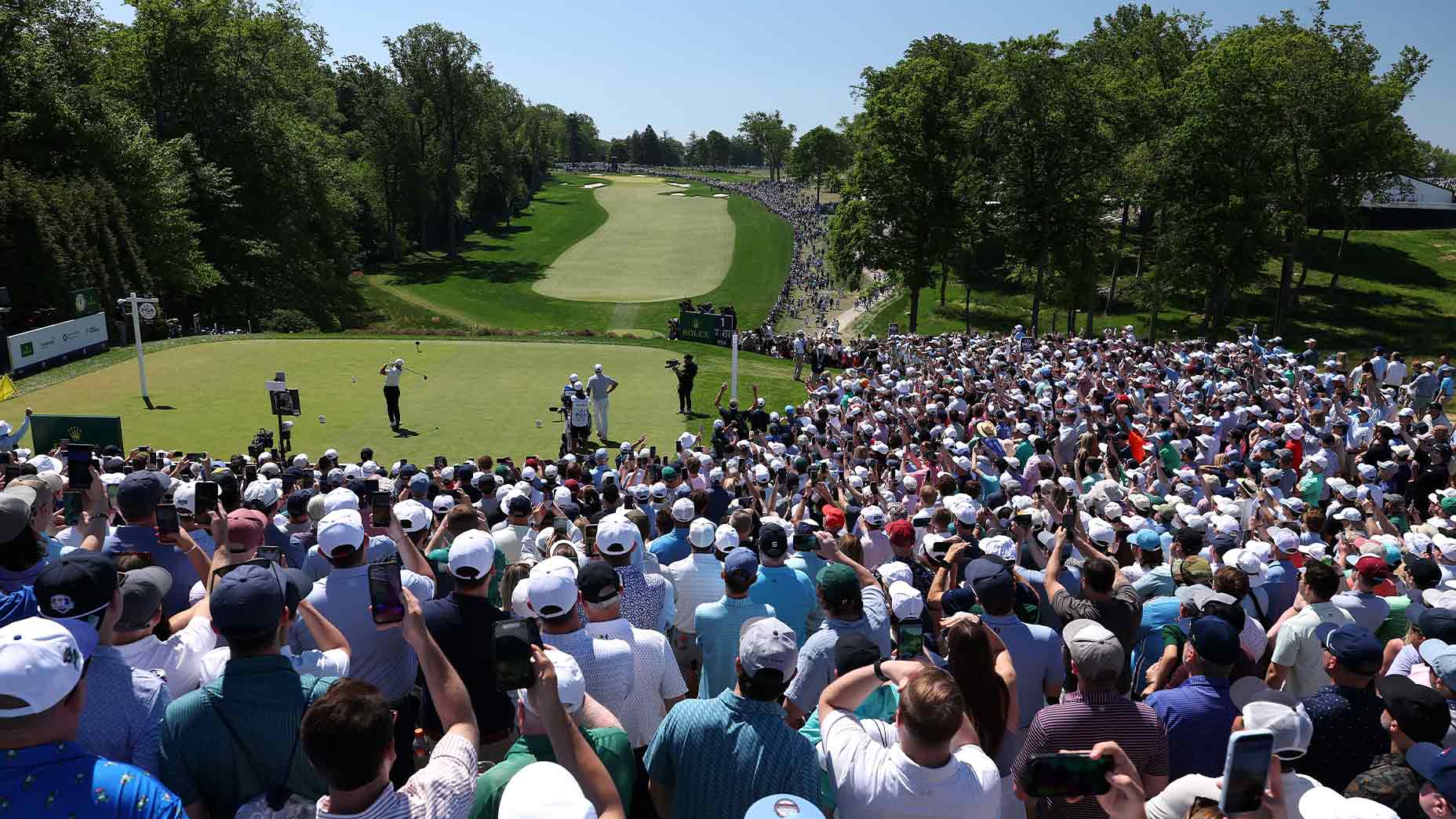 A large crowd watches a golfer tee off at the PGA Championship on a sunny day, with lush trees and a green fairway in the background. Many spectators hold up phones to capture the moment.