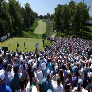 A large crowd watches a golfer tee off at the PGA Championship on a sunny day, with lush trees and a green fairway in the background. Many spectators hold up phones to capture the moment.