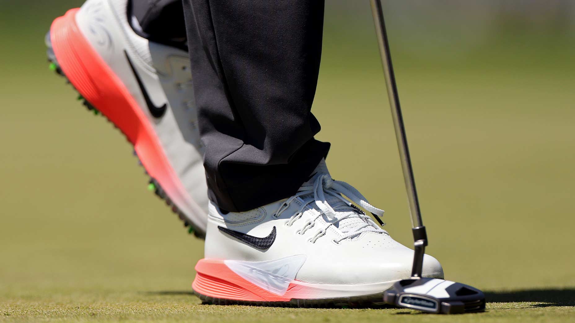 A close-up of Scottie Scheffler&rsquo;s white Nike golf shoes with orange soles, standing on the green at the PGA Championship, preparing to putt. The focus is on the shoes and putter head against the grass.