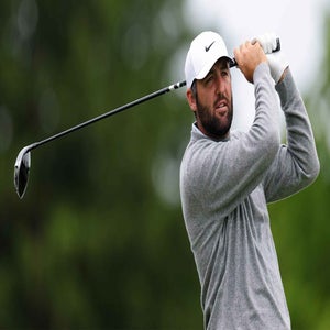 Scottie Scheffler, a PGA Championship favorite, wearing a gray sweater and white cap swings a golf club outdoors, with a blurred green background behind him.