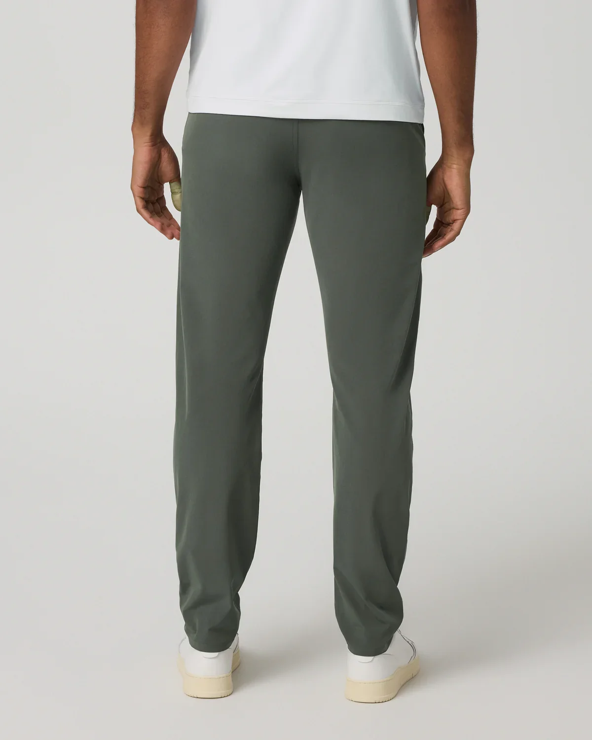 A man wearing dark green pants, a white shirt, and white shoes is standing facing away from the camera with a light background, ready to choose the best golf bags for his next trip.