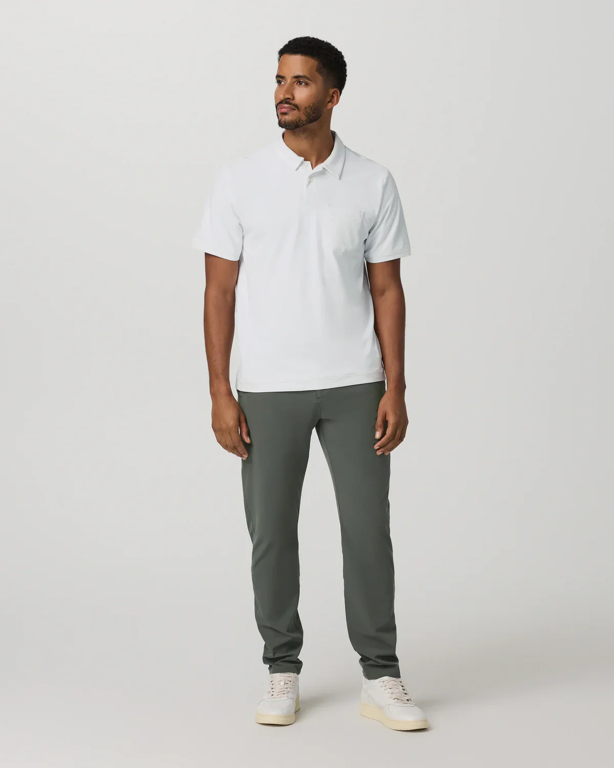A man stands against a plain light background, wearing a white short-sleeve polo shirt, gray pants, and white sneakers—an effortless style ideal for those searching for the best golf travel bags for their next getaway. He looks slightly to the side with a relaxed posture.
